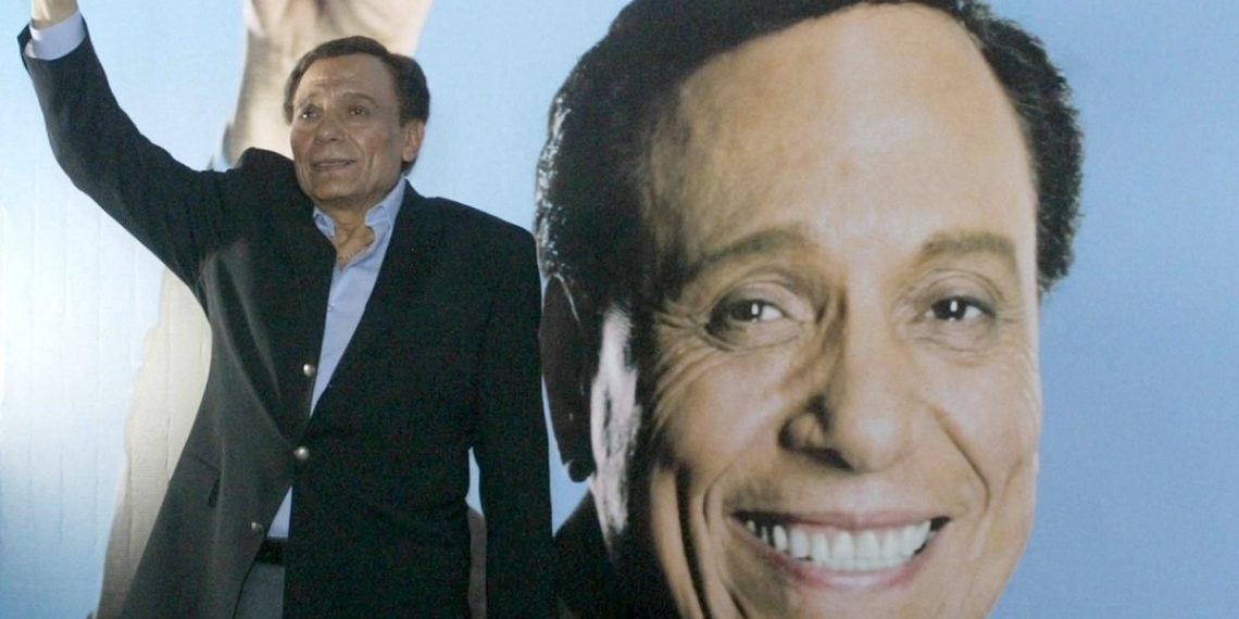Egypt's top comedy star Adel Imam poses in front of a huge self-portrait on a poster advertising his new film Al-Safarah Fil Omarah (The Embassy in the Building) during shooting in Cairo 08 May 2005. The comedy film will be released this summer. AFP PHOTO/AMRO MARAGHI (Photo by AMRO MARAGHI / AFP)